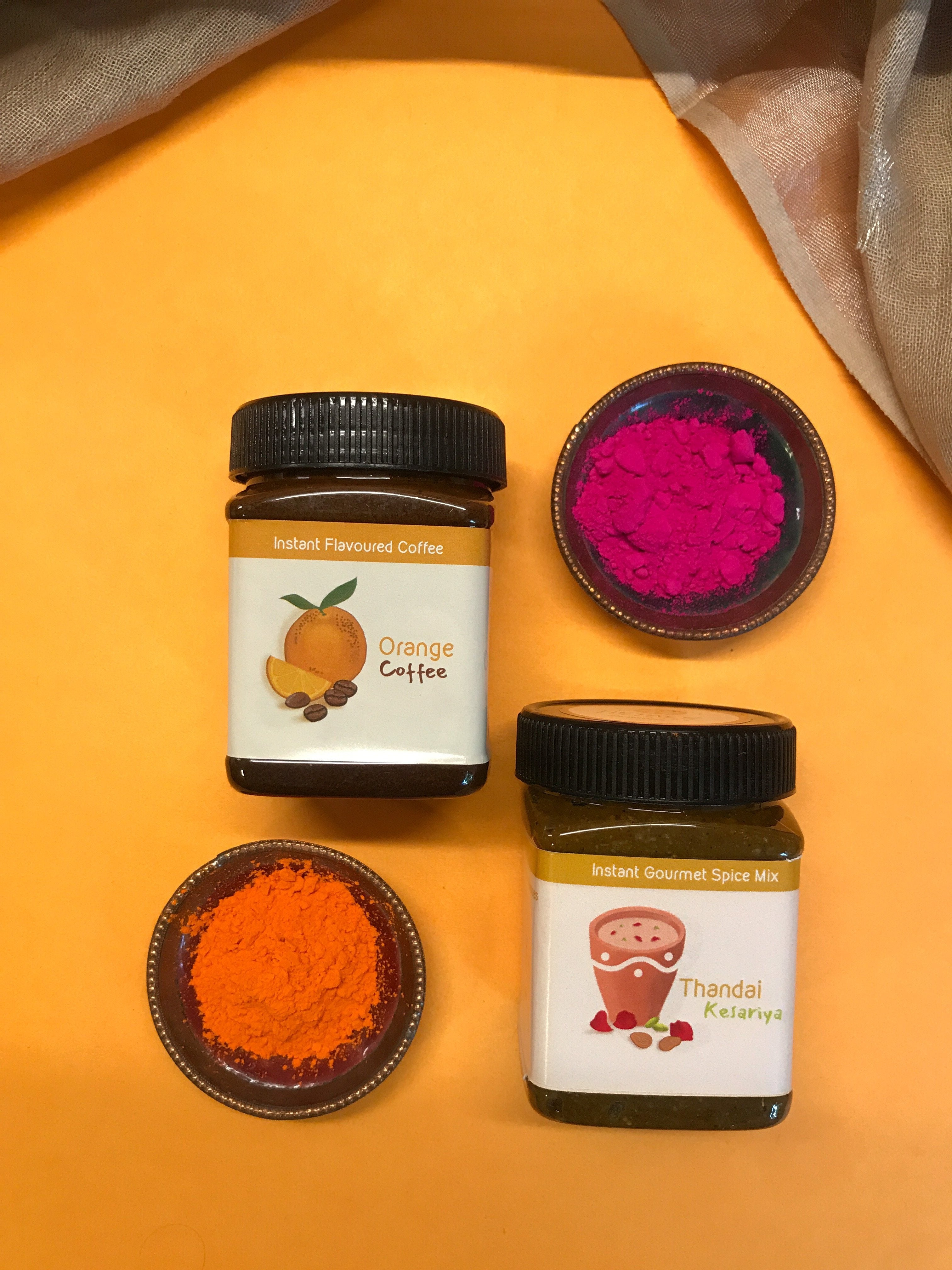 An image of Hetal's Homemade Holi Hamper. The hamper consists of two of our products. Thandai and Orange flavoured instant coffee. The image also shows organic holi colours in a bowl for decorative purposes.